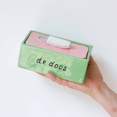 een doos van keramiek met op de deksel een tampon als handvat. De deksel is roze, de doos zelf groen. Op 1 zijkant staat 'de doos' geschreven