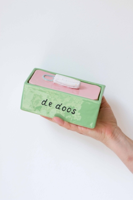 een doos van keramiek met op de deksel een tampon als handvat. De deksel is roze, de doos zelf groen. Op 1 zijkant staat 'de doos' geschreven