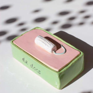 lichtgroen bewaarkistje van keramiek met een roze deksel en als handvat een tampon, op de zijkant staat 'de doos' geschreven